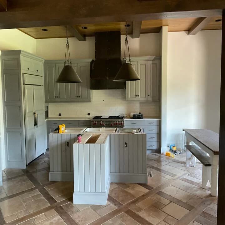 This is an image from Northwind Painting LLC. The image shows a kitchen renovation with newly painted gray cabinetry, matching island panels, and coordinated wall paint enhancing the space’s warm wood beams and tiled flooring.