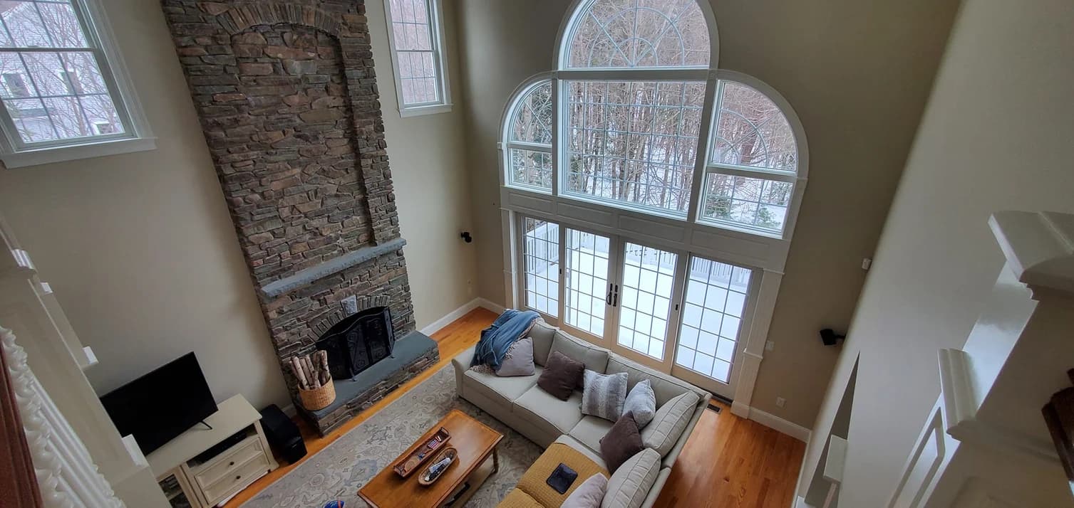This is an image from WC Painting Inc, located in Massachusetts. The interior living room showcases tall windows, a stone fireplace, and freshly painted beige walls with white trim, enhancing the natural light and architectural detail.