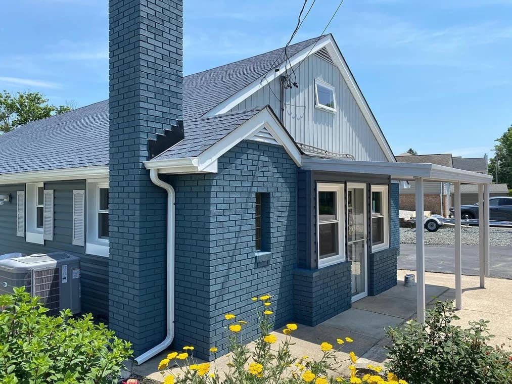 This is an image from Aurora Painting, located in Pennsylvania. The image shows a freshly painted blue brick home with white trim, a covered side entry, and bright yellow flowers in the foreground.