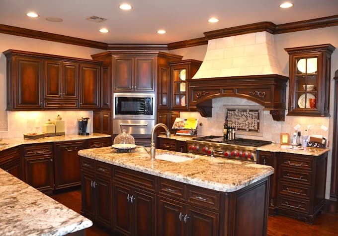 This is an image from JT Paint & Design, located in Oklahoma. The photo shows a traditional kitchen with dark wood cabinetry, granite countertops, a large center island, and an ornate range hood with detailed trim.