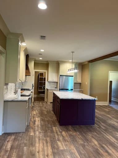 This is an image from Yeras Painting, located in Louisiana. The photo shows a modern kitchen interior with freshly painted cream cabinets, a dark purple island, stainless steel appliances, and wood-look flooring.