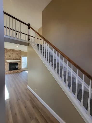 This is an image from Skywalkers Painting & Repairs, located in Ohio. The image shows a freshly painted interior staircase with white balusters, a wood handrail, beige walls, and light hardwood flooring leading to a living area with a fireplace.