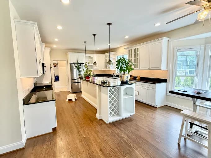 This is an image from Perfect Painters & More, located in Maryland. The photo shows a bright, modern kitchen with freshly painted white cabinets, black countertops, hardwood flooring, and pendant lighting above a central island.