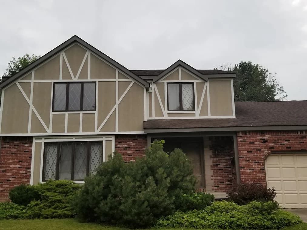 This is an image from Queen City Painting & Wash, located in Ohio. The photo shows a Tudor-style home exterior before repainting, with tan stucco panels, white trim, and partial red brick façade.