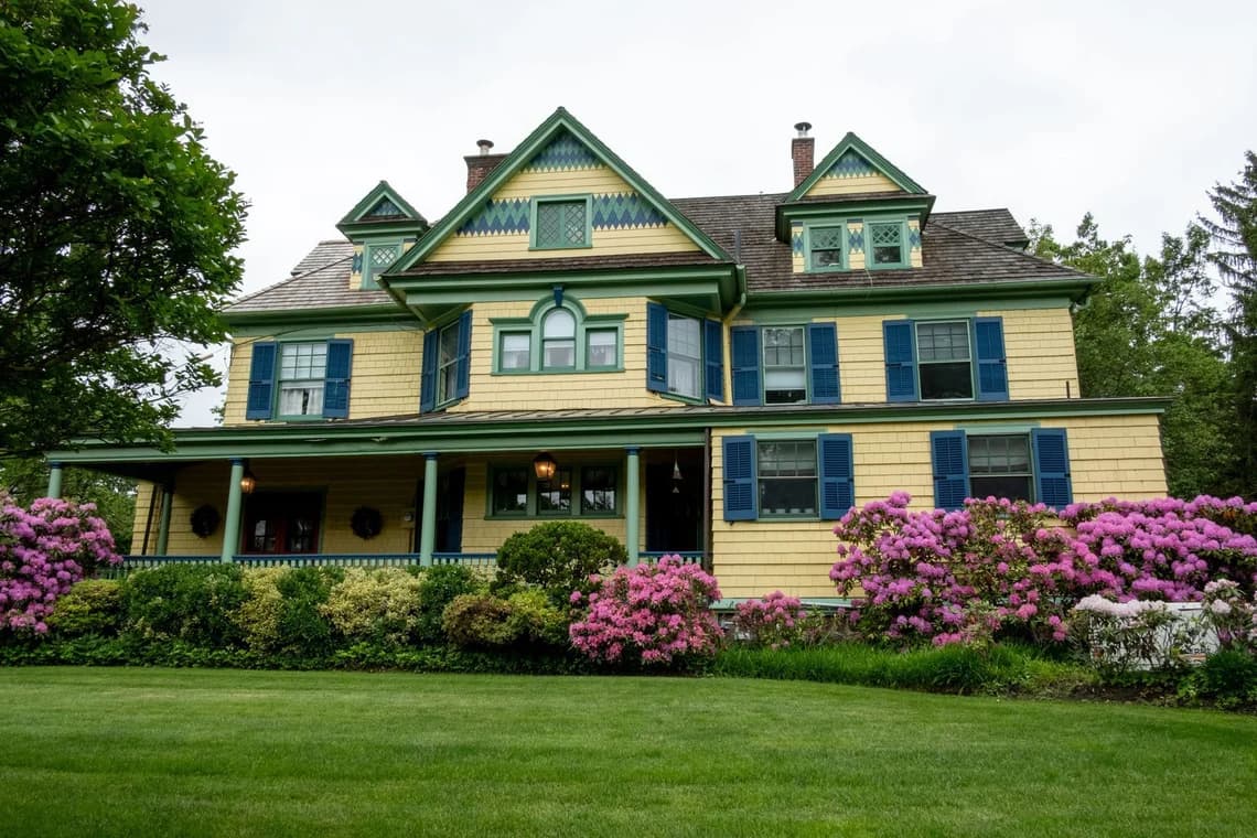 A historic yellow and green Victorian-style home with blue shutters and lush landscaping.