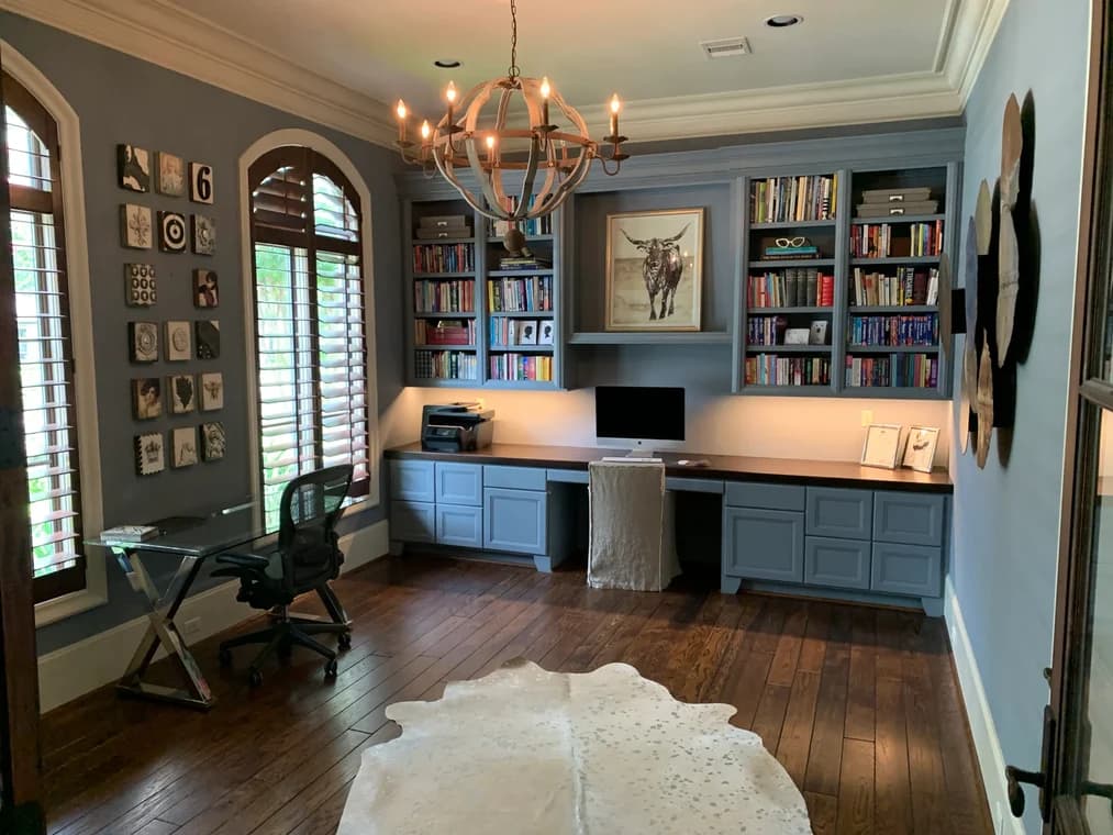 "This is an image from Squared Away Painting, located in Texas. The home office features blue painted built-in shelving, matching cabinetry, and warm wood flooring with decorative wall art and a chandelier."