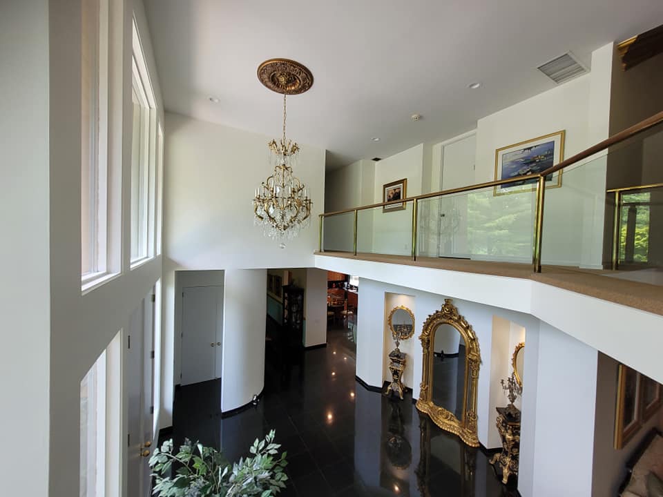 This is an image from JRT Pro Painters, located in New Jersey. The image shows a large interior foyer with freshly painted white walls, high ceilings, a chandelier, balcony railing, and polished dark flooring.