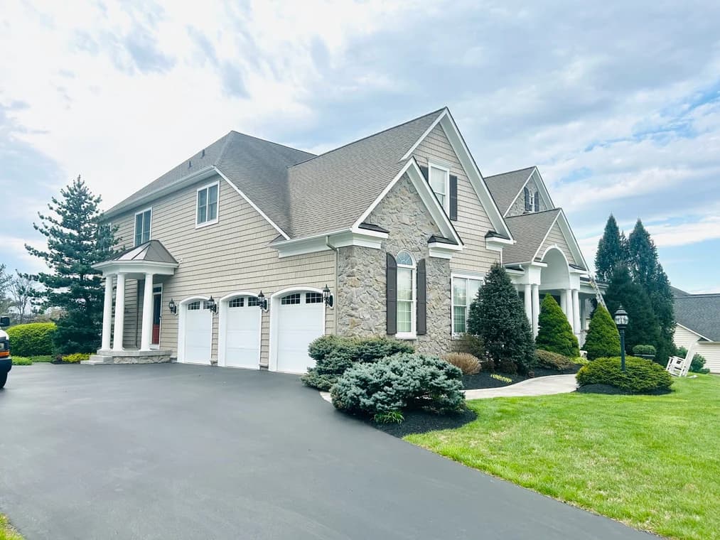 This is an image from Painting Contractors of MD, located in Maryland. The photo shows a large modern home exterior with tan siding, stone accents, white trim, and a freshly coated black asphalt driveway.