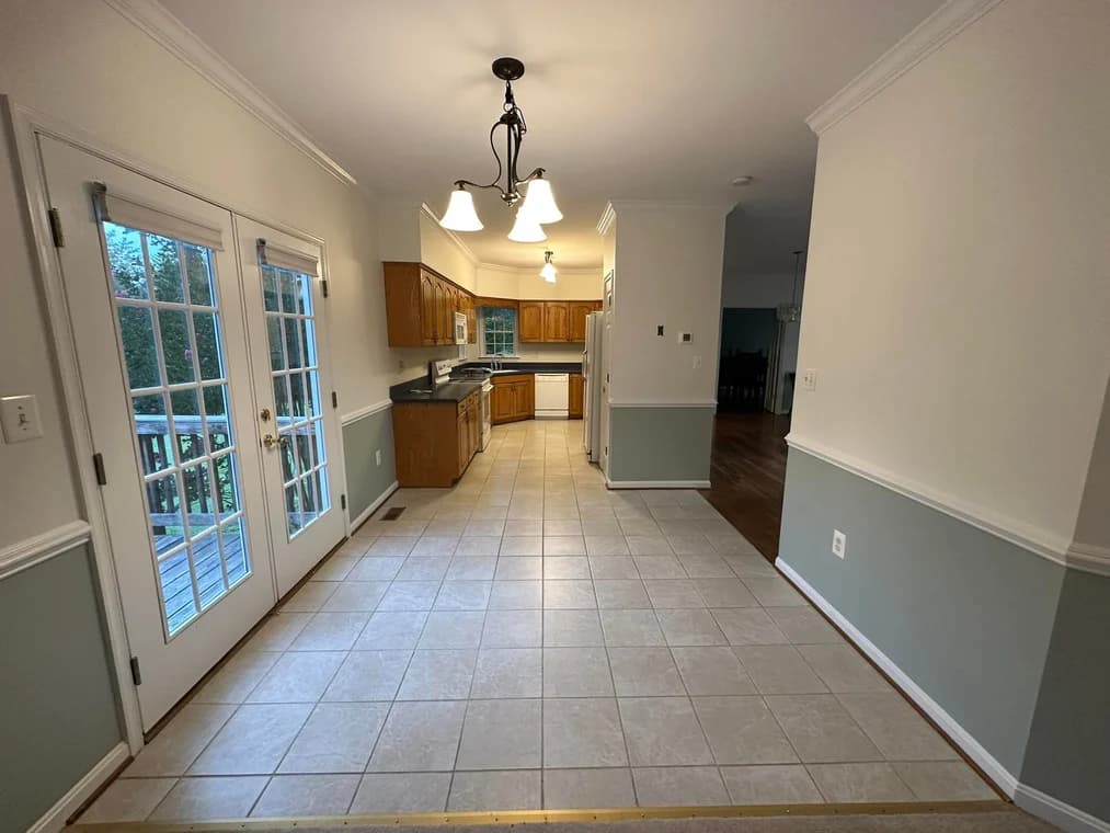 This is an image from DJ’s Painting & Powerwashing, located in Maryland. The image shows a light-filled kitchen with white walls, two-tone wainscoting, tile floors, and French doors opening toward an exterior deck.