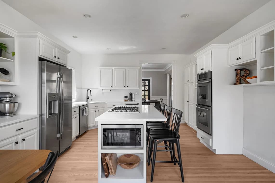 This is an image from AH Painting Services, located in Massachusetts. The photo displays a bright, modern kitchen with white cabinetry, stainless steel appliances, and a central island, emphasizing the company’s expertise in clean, high-quality interior painting.
