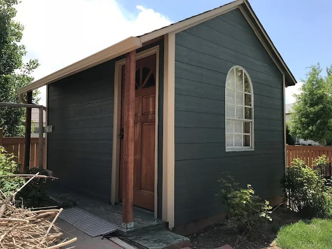 This is an image from Noble Painting company, located in Fort Collins, CO The image is a small detached shed or studio with dark green painted exterior, arched window, natural wood front door, and tan trim, exterior repainting by Noble Painting