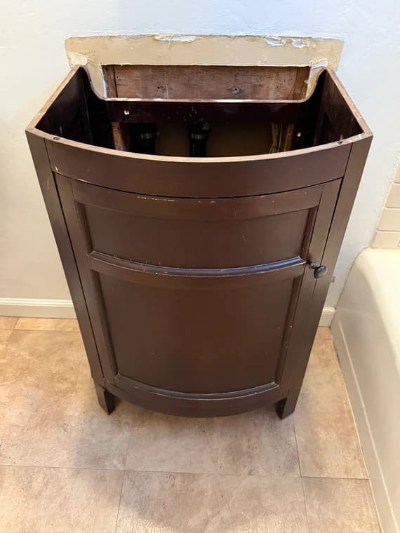 Dark wood vanity base with top removed, exposing interior plumbing and wall damage before update