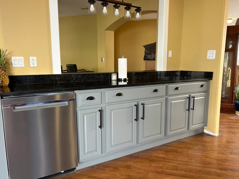 This is an image from CertaPro Painters of Albuquerque, located in New Mexico. The photo shows freshly painted gray kitchen cabinets with black hardware, complemented by a black granite countertop and stainless steel dishwasher.