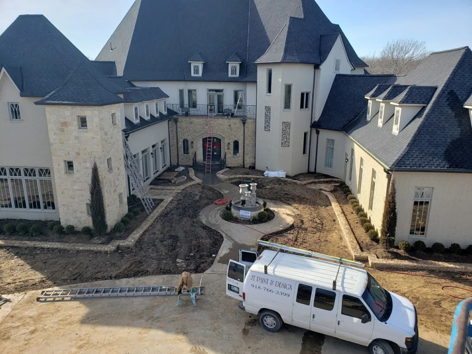 This is an image from JT Paint & Design, located in Oklahoma. The photo shows a large residential property under renovation with a JT Paint & Design work van parked in front, ladders set up, and painting equipment visible on site.