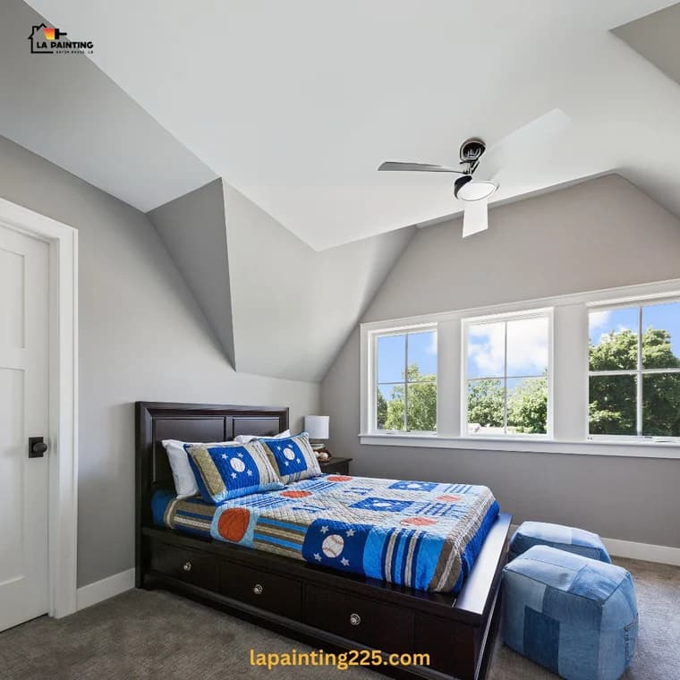 This is an image from LA Painting, located in Louisiana. The image shows a freshly painted bedroom with soft gray walls, white trim, and a sloped ceiling, featuring a dark wood bed frame with a blue sports-themed quilt and large windows letting in natural light.