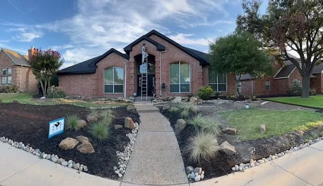 This is an image from Platinum Painting of Richardson, located in Texas. The exterior image displays a brick home with updated trim and entryway painting, viewed from a walkway lined with landscaping stones and plants.