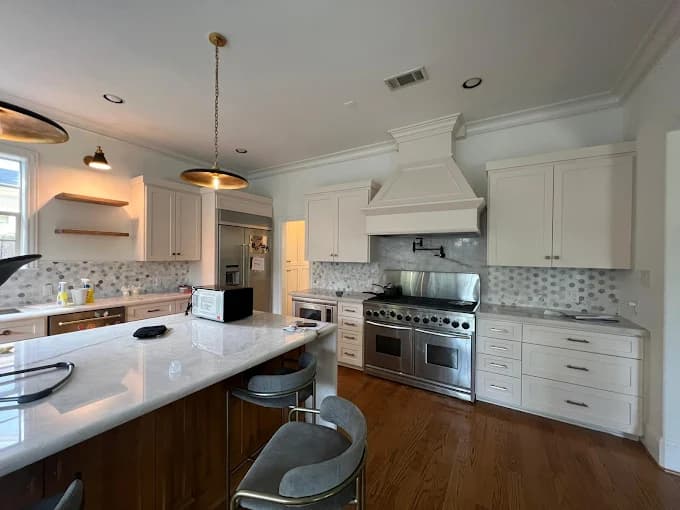 This is an image from Blue Painting, located in Texas. The image shows a bright kitchen with white cabinetry, a large stainless steel range and hood, marble countertops, wood flooring, and gold pendant lighting above a spacious island.