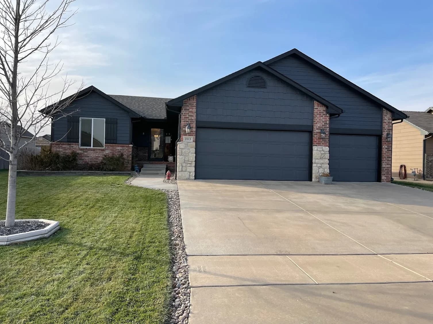 This is an image from Northwind Painting LLC. The image shows a dark charcoal exterior repaint on a single-story home with brick and stone accents, including freshly painted siding, trim, shutters, and garage doors.