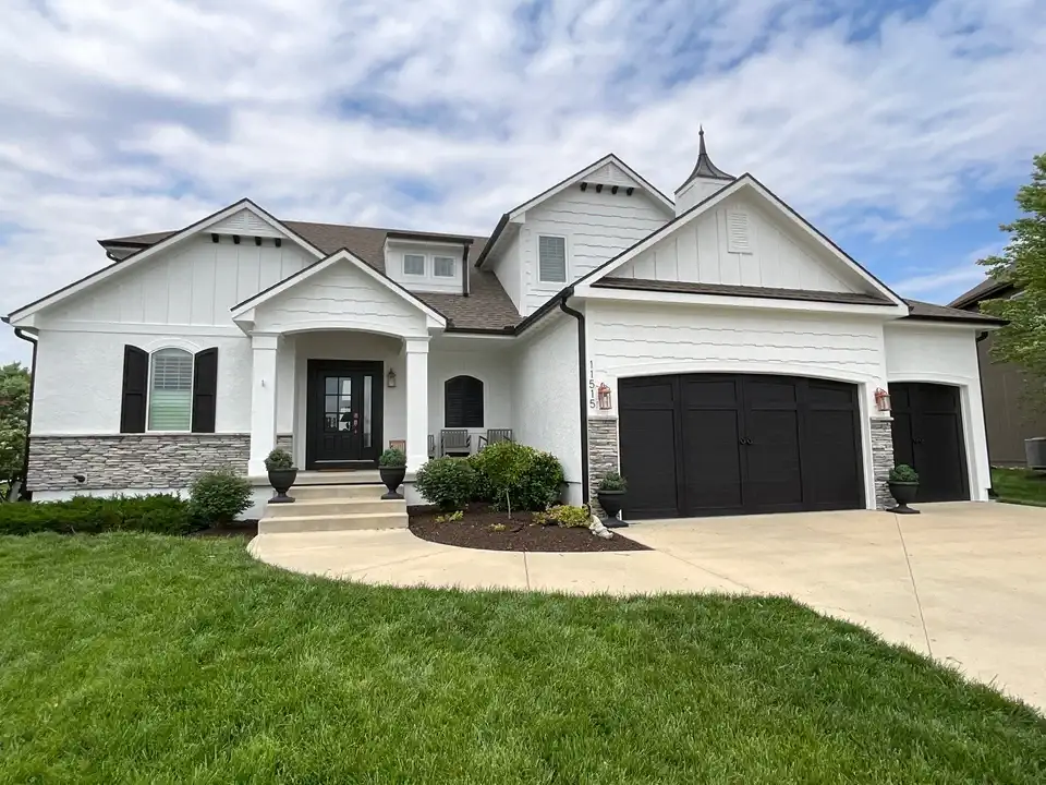 This is an image from PEMCO Painting Interior & Exterior Painter Overland Park.The image is an exterior home in Overland Park KS with fresh white siding, stone accents, and newly painted black shutters and garage doors.