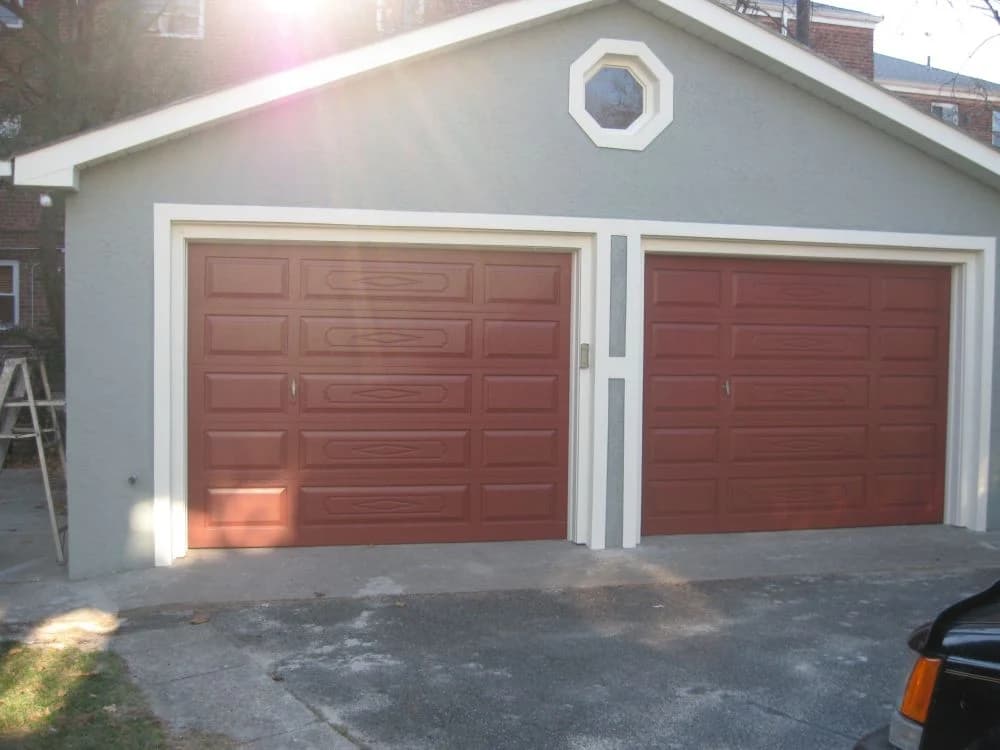 This is an image from JNG Painting LLC. It features a double garage with gray stucco walls and freshly painted red doors outlined in white trim.