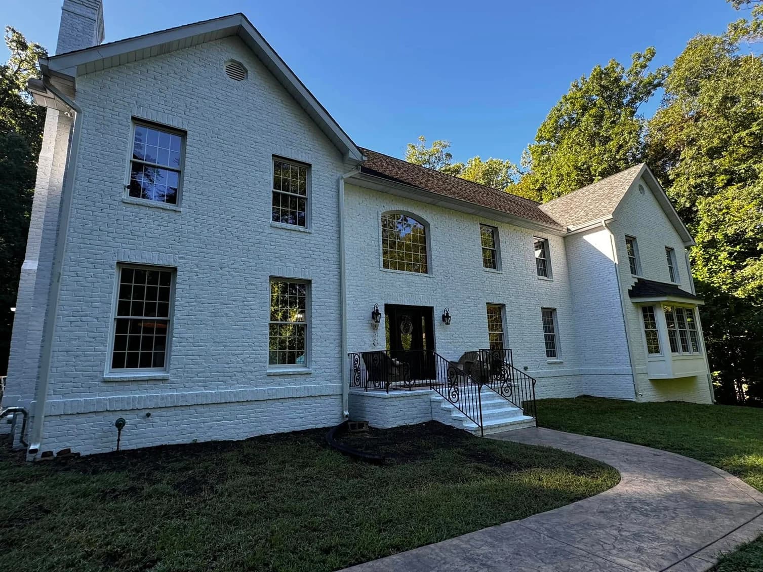 This is an image from Goode Design Painting & Wallcovering Inc., located in Virginia. The photo shows a large two-story home with freshly painted white brick exterior surrounded by lush green trees