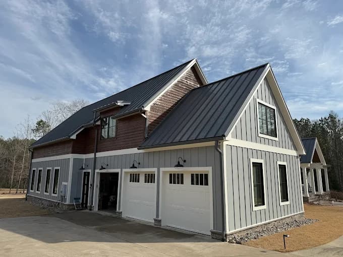 This is an image from Gonzalez Painters & Contractors, Inc., located in North Carolina. The photo shows a freshly painted modern farmhouse-style home with a dark metal roof, gray siding, white trim, and double garage doors under a clear sky.