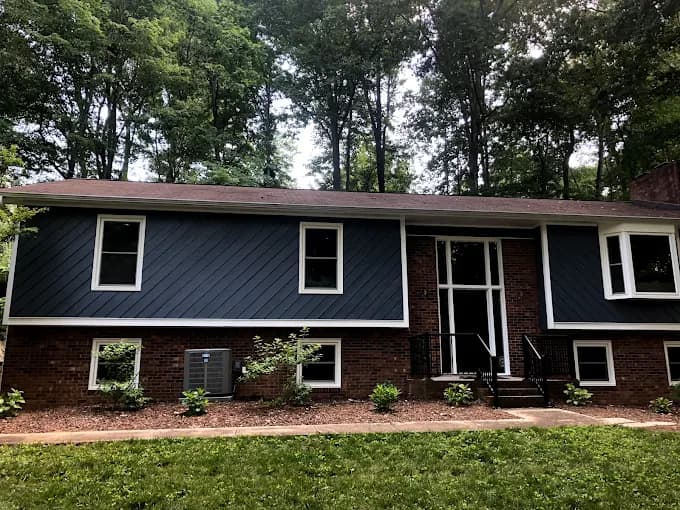 Exterior painting project by Still Perfection Painting Co. in Belews Creek, North Carolina. The image shows blue siding, white trim, trees, and brick skirting.