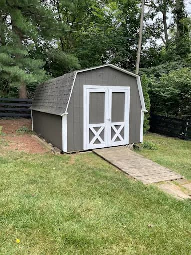 This is an image from R.E.A.L. Painting, located in Kentucky. The image shows a small gray shed with white trim and double barn-style doors surrounded by a green yard.