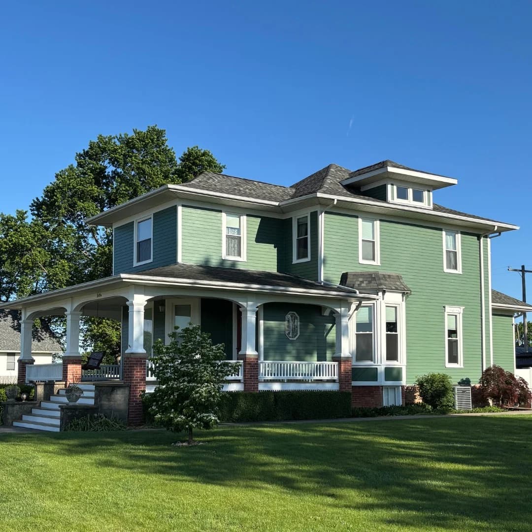 Exterior painting project by Clean Cut Painting & Handyman in Decatur, Illinois. The image shows a green two-story home with white trim, a front porch, and brick columns.