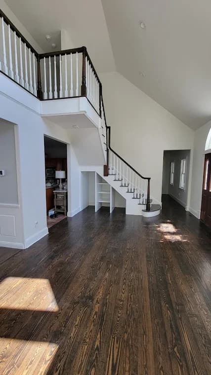 This is an image from CRD Painting Inc, located in New York. The image shows a large open living space with refinished dark hardwood floors, white walls, and a staircase with dark handrails.