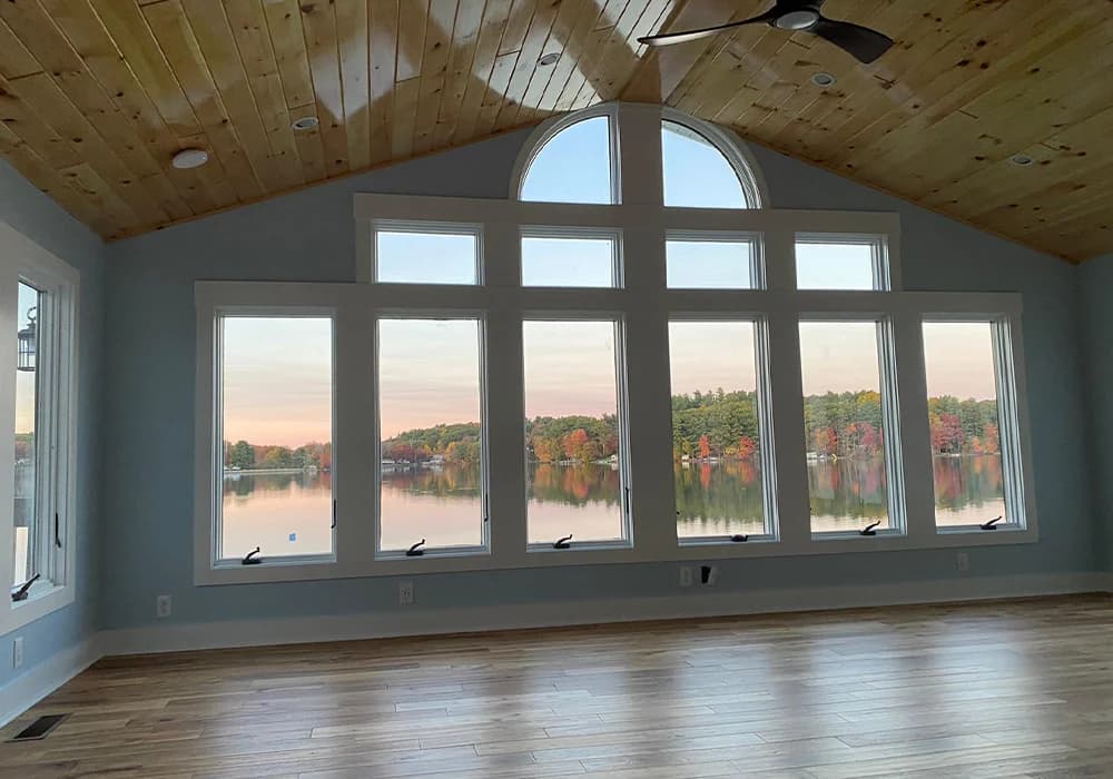 This is an image from Zuck Painting, located in Massachusetts. It highlights a large living space with pale blue walls, expansive windows overlooking a lake, and a wooden ceiling.