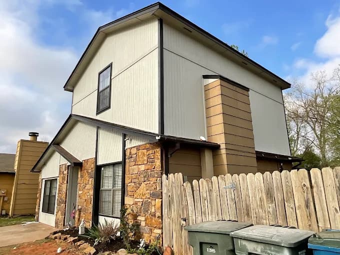 This is an image from CertaPro Painters of OKC, located in Oklahoma. The photo shows a two-story home exterior with freshly painted cream siding, tan accents, dark trim, and natural stone details on the lower level.