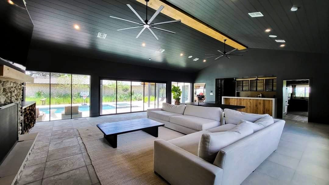 "This is an image from Reyes Painting Co., located in Texas. The interior scene showcases a modern living area with dark walls and ceiling, large sliding glass doors, oversized ceiling fans, and white sectional sofas facing a pool."