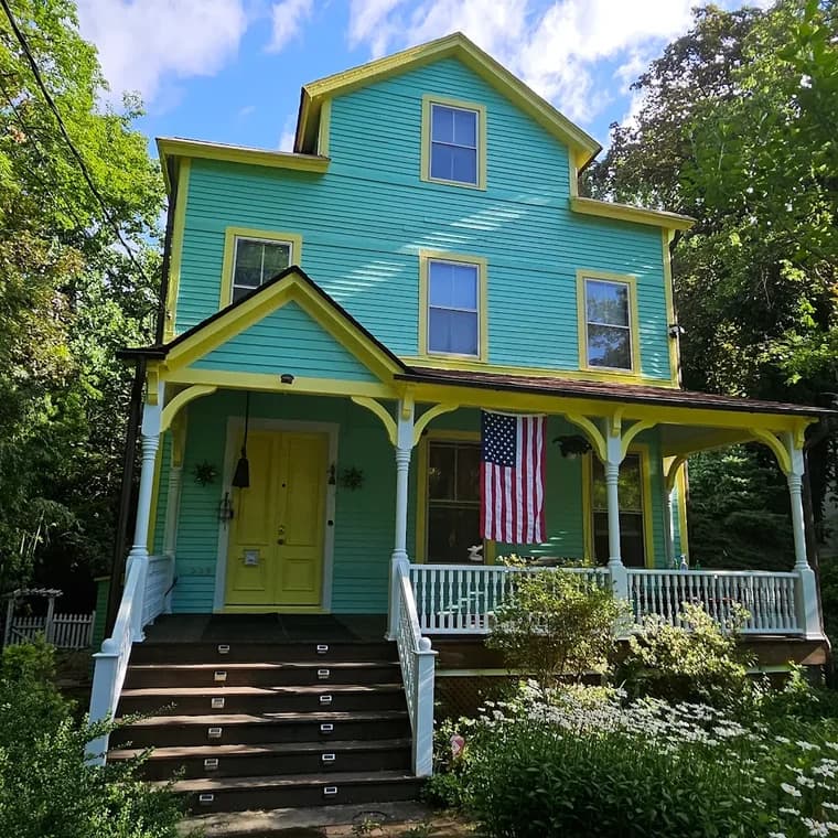 This is an image from CertaPro Painters of Staten Island, located in New York. The photo shows a freshly painted turquoise and yellow exterior of a multi-story home with detailed trim, a covered porch, and an American flag hanging out front.