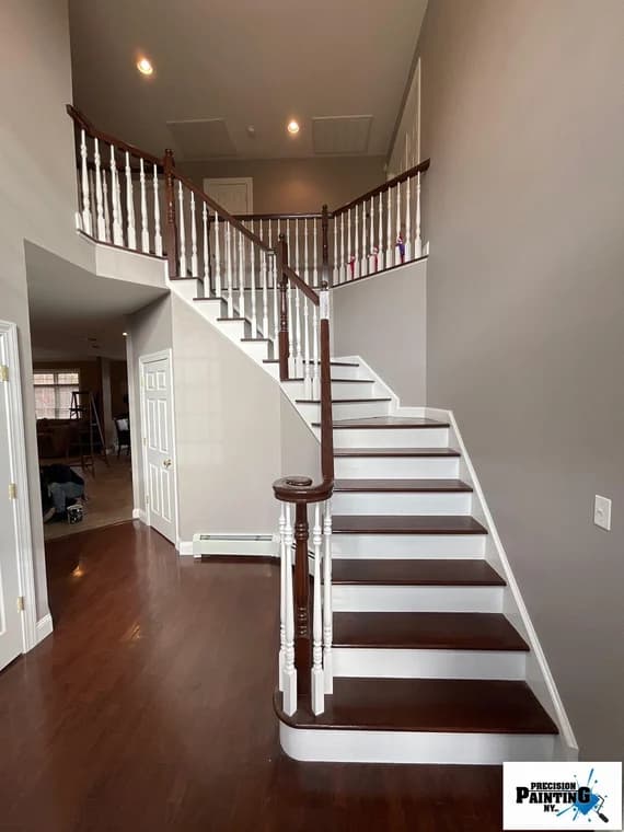 This is an image from Precision Painting NY, located in New York. The image shows a freshly painted interior staircase with gray walls, white balusters, and dark wood steps and handrails.