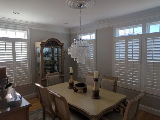 This is an image from CertaPro Painters of Columbia, located in Maryland. The image shows a bright dining room with beige walls, white trim, plantation shutters, and an elegant crystal chandelier above a dining table.