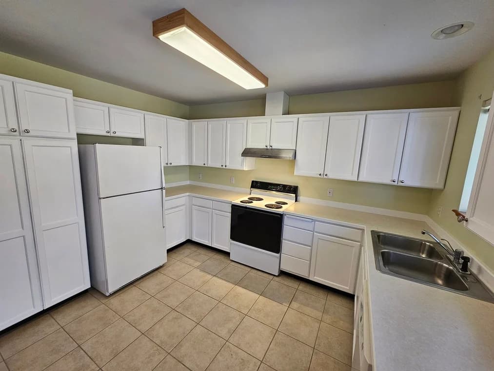 This is an image from Honey Do Painting LLC company, located in Bonners Ferry, ID The image is a clean and updated kitchen with freshly painted white cabinets, light green walls, beige tile flooring, white appliances, and overhead fluorescent lighting, refinishing by Honey Do Painting