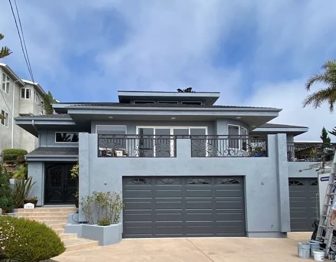 This is an image from Browder Painting Company, located in California. The image shows a newly painted modern two-story home exterior with smooth gray stucco walls, a dark gray garage door, metal balcony railing, and a polished contemporary appearance.