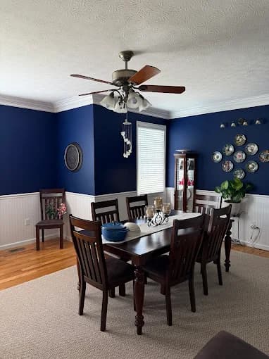 This is an image from Mendez Drywall & Painting LLC, located in Maryland. The image shows a dining room with deep blue painted walls, white wainscoting, a dark wood table set, and hardwood flooring.
