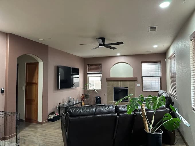 This is an image from New Mexico's Painting Specialists, located in New Mexico. The image shows a freshly painted living room with soft mauve walls, a modern ceiling fan, a mounted TV, a fireplace, and brown leather furniture.