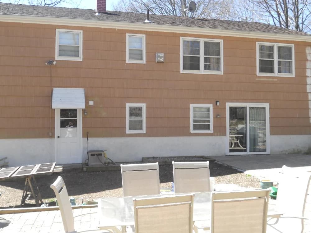 Rear view of a house with brown siding and white trim, featuring multiple windows and a sliding glass door opening to a patio with chairs.