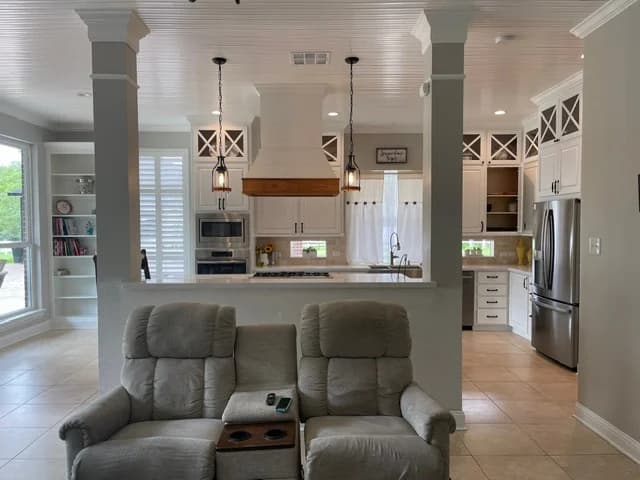 "This is an image from All Seasons Painting LLC, located in Texas. The image shows a white kitchen viewed from a living room, with pendant lights, a custom vent hood, built-in shelving, and stainless steel appliances."