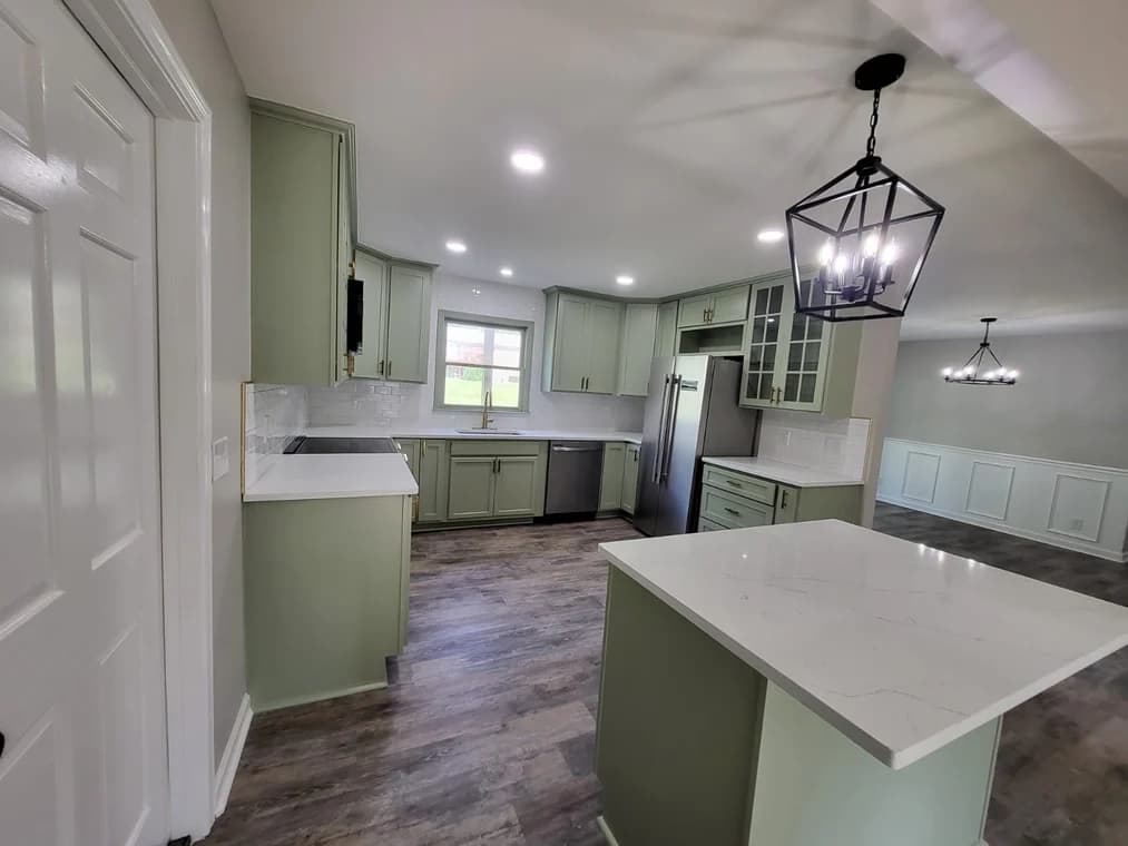 This is an image from Gomez Painting LLC, located in Kentucky. The kitchen features newly painted sage green cabinets, white quartz countertops, and modern black lighting fixtures for a refined, updated look.