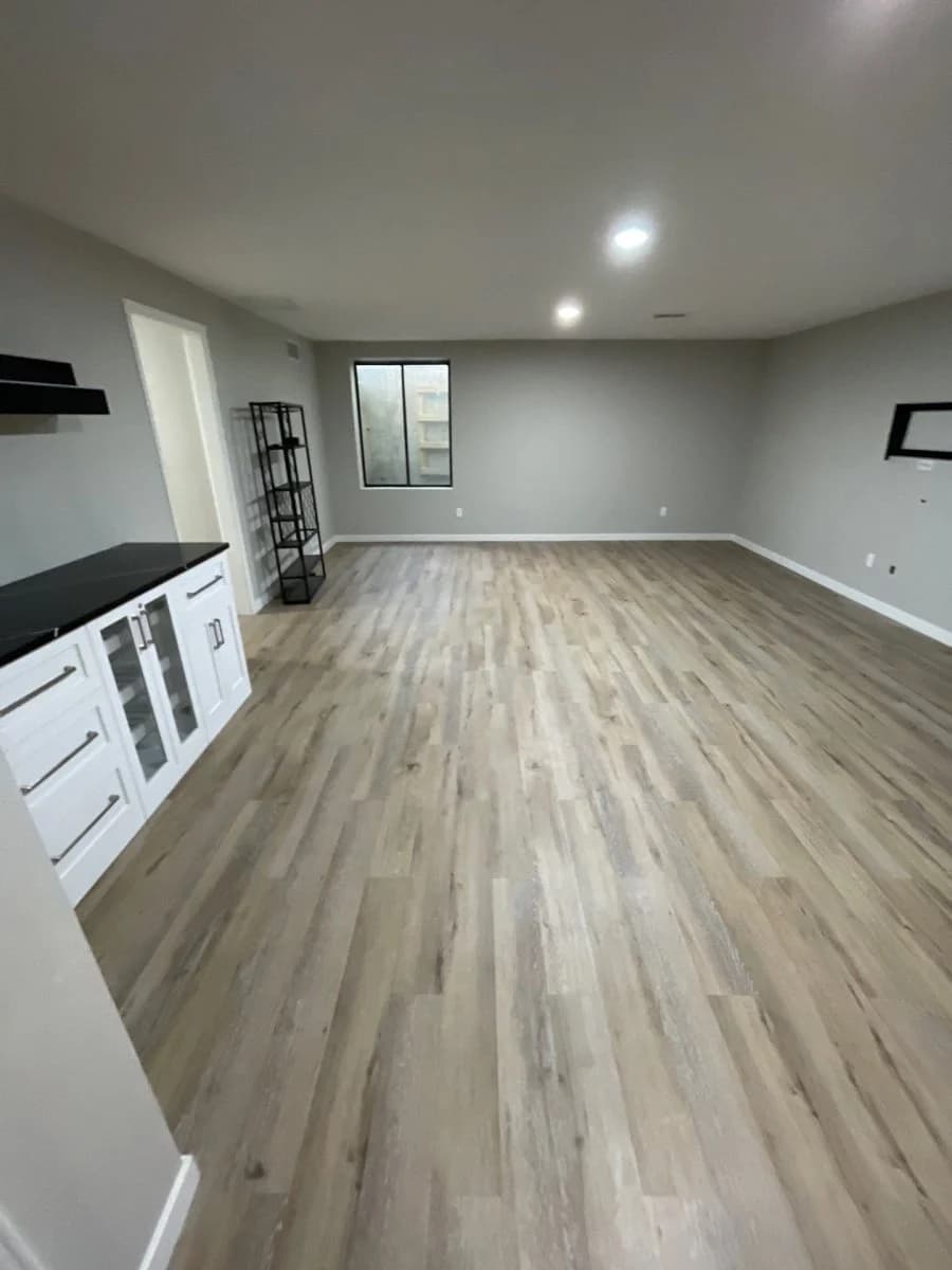 This is an image from River City Painting Inc. The image shows a finished basement with gray painted walls, recessed lighting, and wood-look flooring.