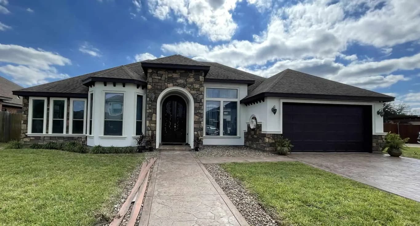 "This is an image from Armored Painting, located in Texas. The exterior home photo displays white stucco accents, stone masonry, dark trim, and a freshly coated garage door under a bright sky."