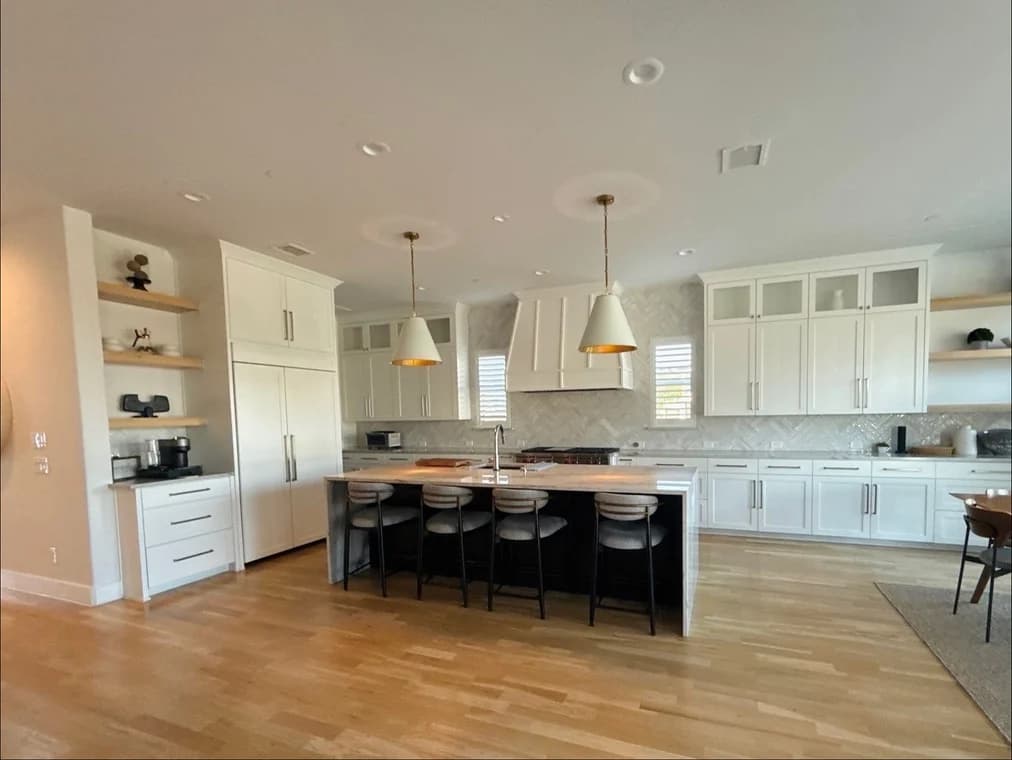 This is an image from Spray Tex Painting, located in Texas. The photo shows a bright modern kitchen with white cabinetry, a large island with barstools, pendant lighting, and hardwood floors.