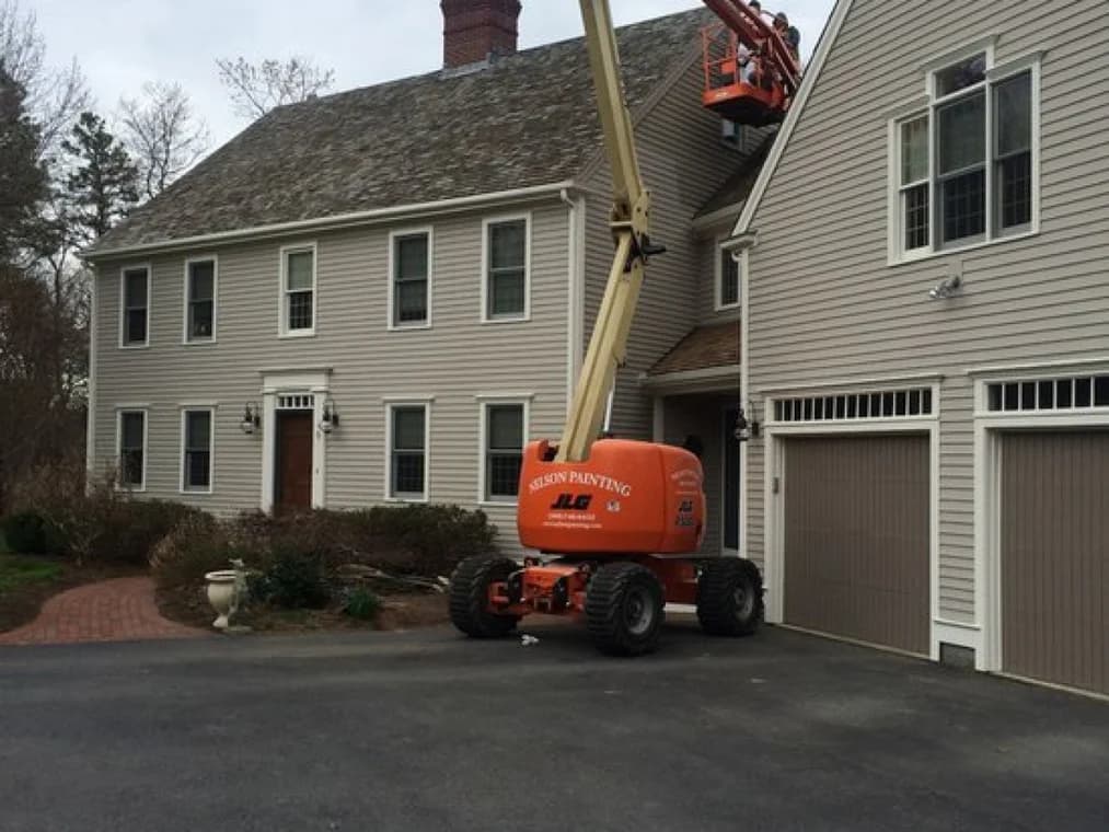 This is an image from Nelson Painting, Inc., located in Massachusetts. It shows exterior painting work on a large beige home with a lift being used to reach the upper sections of the siding.