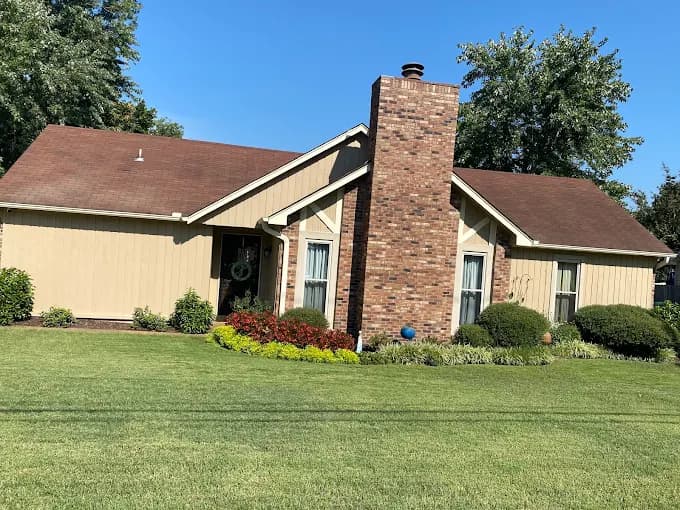 Exterior painting by Riggins Painting in Bartlett, Tennessee. The image shows a tan single-story home with a brick chimney, dark trim, and green lawn.