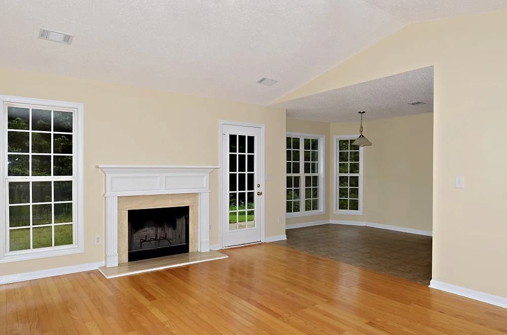 This is an image from Promax Painters company, located in Boynton Beach, FL. The image is a freshly painted living room and dining area with cream-colored walls, hardwood floors, fireplace, and large white-trimmed windows