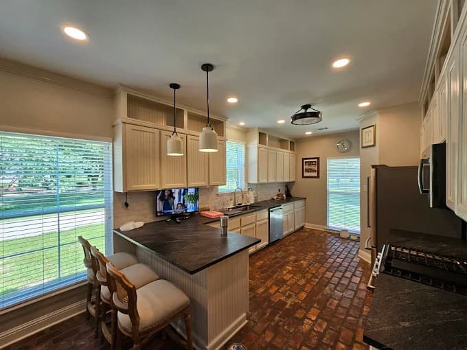 This is an image from Southern Dropcloth Painting LLC, located in Louisiana. The image shows a renovated kitchen with cream-colored cabinets, dark countertops, pendant lighting, and a rustic red brick floor creating a warm and inviting atmosphere.